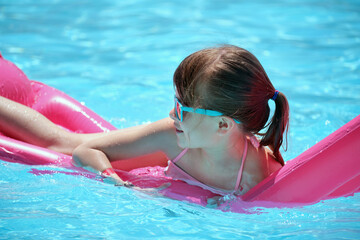 Happy child girl relaxing on inflatable air mattress in swimming pool on sunny summer day during tropical vacations. Summertime activities concept