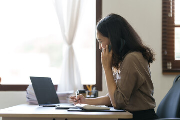 Happy asian businesswoman sitting at desk talking with mobile phone with somebody in office workplace.