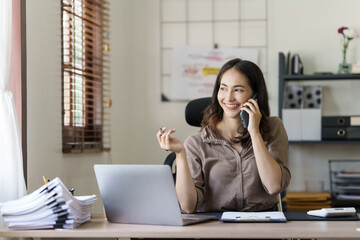 Happy asian businesswoman sitting at desk talking with mobile phone with somebody in office workplace.