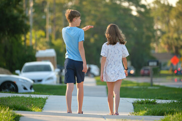 Obraz premium Rear view of two young teenage children, girl and boy, brother and sister walking together on rural street on bright sunny day. Vacation time concept