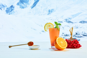 Glass with orange tea or cocktail, berries and honey on white table over background of landscape of winter snow-capped mountains. Health, care, love