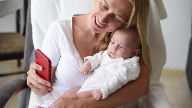 Mom holds her newborn baby and talk by video call with father by phone. child look on screen of phone. happy family smile mother and kid. online life 
