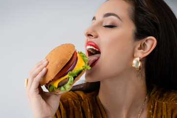 elegant young woman with makeup eating delicious burger isolated on grey.