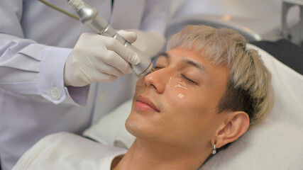 Fototapeta na wymiar A handsome Asian man undergoes facial rejuvenation in a beauty clinic using modern medical equipment. By certified beauty experts according to standards.