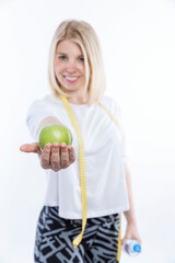 Woman with a green apple and a bottle of water in her hands. Pretty smiling blonde woman in a white tank top with a measuring tape around her neck. White background. Vertical.