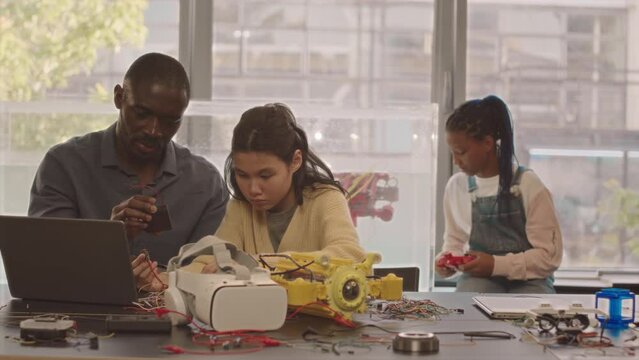 Medium slowmo of African American teacher of electronics and group of his smart diverse students experimenting with robo boat and other electronic devices in school laboratory