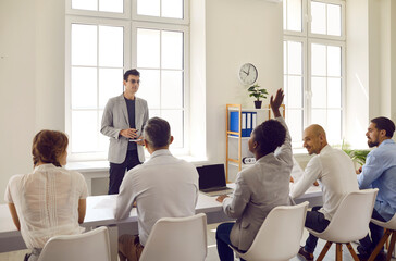 Team of diverse people having class with a professional business coach. Group of multiracial male and female corporate employees sitting at office desks and asking questions. Business training concept - obrazy, fototapety, plakaty