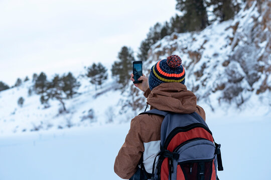 Rear view young man in brown warm clothes with backpack walking in snow among rocks and cliffs in winter looking at view taking photos with smartphone Active lifestyle hiking