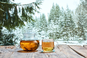 Winter still life. Glass pot with herbal tea on wooden table over winter snow forest background. Design for greeting card, wallpaper for device screen