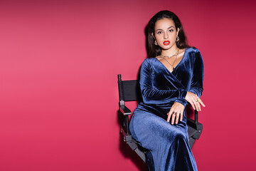 young brunette woman in blue velvet dress looking at camera while sitting on red background.