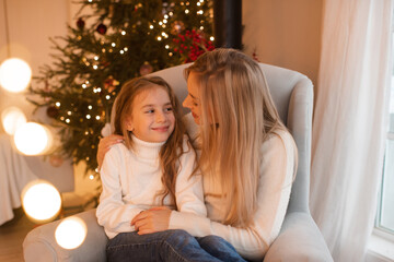 Obraz na płótnie Canvas Mother and kid girl daughter celebrating christmas holidays sitting in chair over xmas tree in room. Winter greeting season. Celebration. Motherhood.