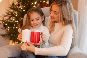 Mother and kid girl daughter celebrating christmas holidays drinking hot chocolate sit in chair over xmas tree lights in room. Winter greeting season. Motherhood. Celebration.