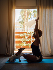 Adorable dog touchingly and faithfully watching the owner young woman doing morning exercises. sitting on the floor on a yoga mat in front of a window in at home in living room. morning affirmations