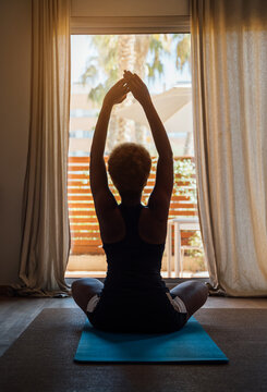 affirmations stretching exercises at the morning. Woman sits in sportswear in front of the window with her hands up. Good morning exercise silhouette with afro hairstyle