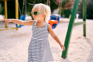 Little girl in sunglasses stands on the playground and points her finger somewhere. High quality photo