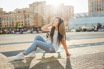 A young hipster girl wearing headphones, sitting on a skateboard, spends time in the city at sunset. evening autumn sunset. generation z