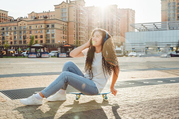Fototapeta na wymiar A young hipster girl wearing headphones, sitting on a skateboard, spends time in the city at sunset. evening autumn sunset. generation z