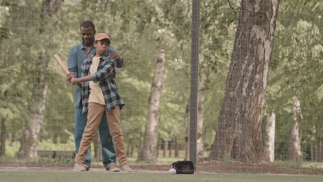 Full length shot of African American man teaching his 12 year old son to bat while practicing baseball outdoors in summer