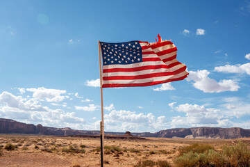 The flag of the United States of America flies in the sun in front of a barren plain in Monument Valley