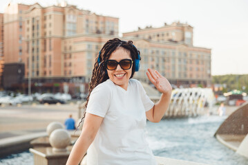 A young cheerful happy woman with dreadlocks, dressed in a white T-shirt, dancing, listening to music with headphones, resting, relaxing in a city park, walking along an alley. Urban lifestyle concept