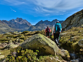 Fototapeta premium Alpine mountain landscape with couple, man and woman, trekking or hiking behind a stone with blue sky with clouds.