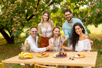 Group of happy young people cheering with fresh lemonade and eating fruits in the garden