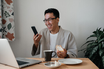 Adult asian man wearing glasses and robe sitting in kitchen using laptop and smartphone. Leisure morning time. Remote job, freelance working concept.