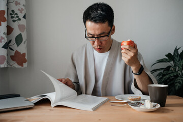Adult asian man wearing glasses reading book or magazine while having breakfast. Leisure morning...