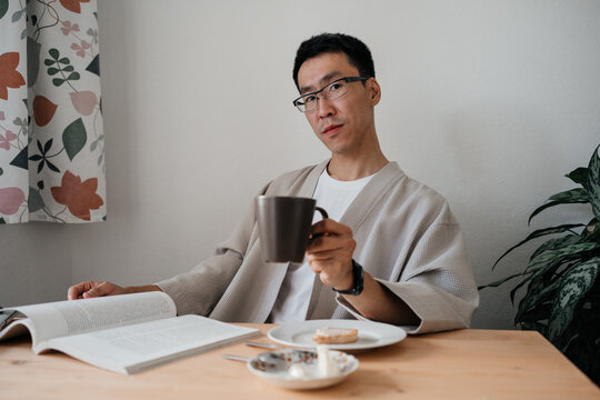 Adult asian man wearing glasses looking at camera reading book or magazine during breakfast. Leisure morning time.