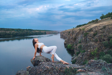 Naklejka na ściany i meble The girl is engaged in yoga, stands in a plank position on a rocky river bank.