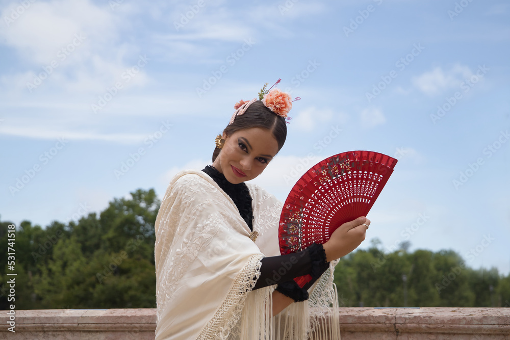 Wall mural Portrait of young teenage girl in black dance dress, white shawl and pink carnations in her hair, dancing flamenco with a red fan. Concept of flamenco, dance, art, typical Spanish dance, fan.