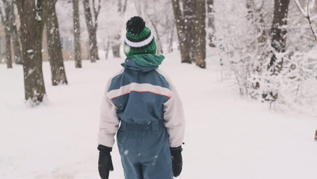 Boy in winter clothes and knitted hat slowly walks through snow-covered park during snowfall. Back view.