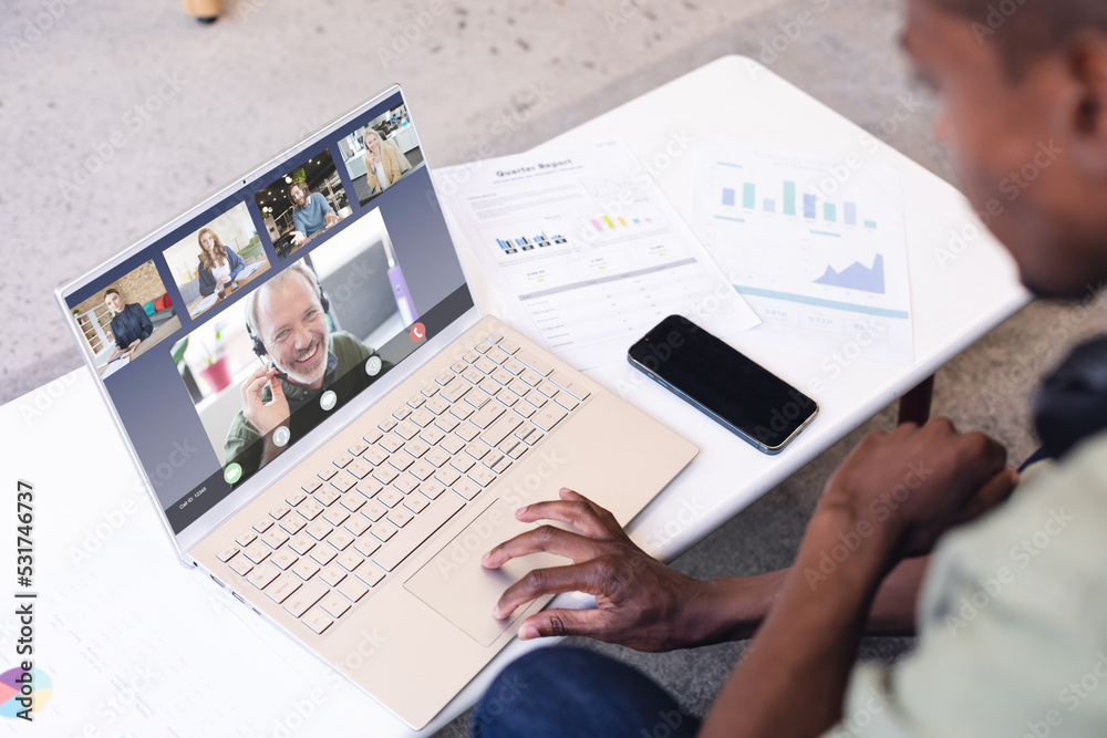 Wall mural Multiracial man with cellphone and documents on desk discussing with coworkers on video call