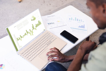 African american male professional with documents and cellphone on desk analyzing report over laptop