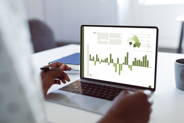 Cropped hands of african american businesswoman holding pen and analyzing graphs over laptop