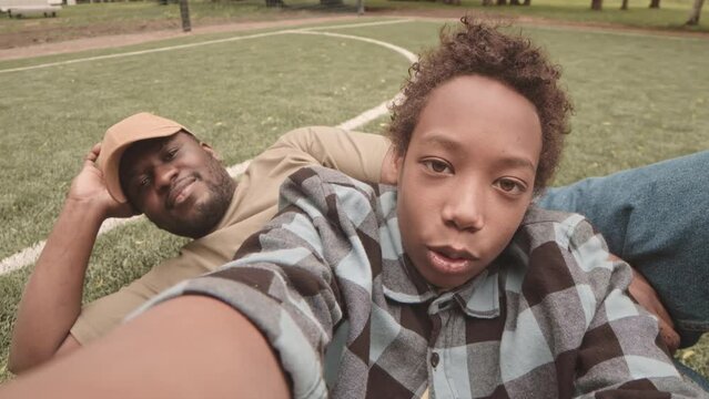 Handheld POV slowmo of African American teenage boy and his dad resting together on green grass at sports field outdoors in park on warm summer day, waving and smiling at camera