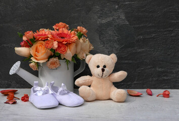 Teddybär mit Blumen und Babyschuhen vor einer dunklen Wand.