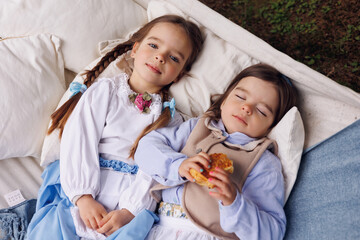 Two little girls laying on hammock in park and eating pretzel and buns. Little baby sisters of 3 and 5 in retro vintage dress having fun and smiling in forest outdoors.