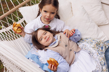 Two little girls laying on hammock in park and eating pretzel and buns. Little baby sisters of 3 and 5 in retro vintage dress having fun and smiling in forest outdoors.