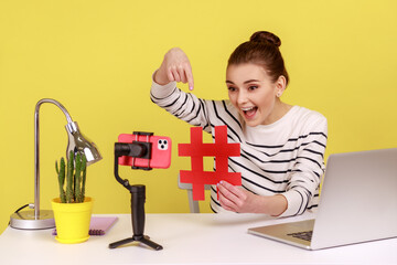 Smiling excited woman blogger showing big red hashtag symbol at smartphone camera, sharing viral...