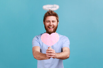 Portrait of bearded angelic man with holy nimbus holding in hands pink paper heart on stick,...