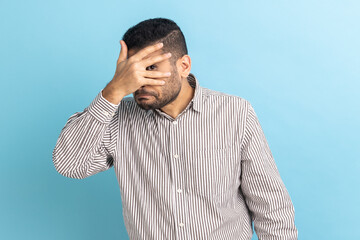 Bearded handsome businessman looking through fingers, peeking with curious face, having suspicious, watching secrets, wearing striped shirt. Indoor studio shot isolated on blue background.