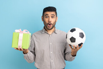 Portrait of shocked surprised businessman standing looking at camera with amazement, holding soccer ball and present box, wearing striped shirt. Indoor studio shot isolated on blue background.