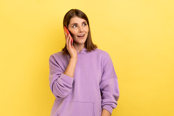 Fototapeta premium Portrait of cheerful woman talking with friend on cell phone, looking away and smiling joyfully, having pleasant conversation, wearing purple hoodie. Indoor studio shot isolated on yellow background.