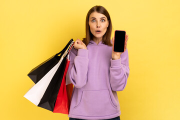 Fototapeta na wymiar Amazed surprised woman standing with excited look showing shopping bags and smart phone with blank screen for advertisement, wearing purple hoodie. Indoor studio shot isolated on yellow background.