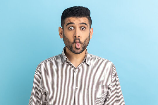 Portrait of funny childish bearded businessman making fish face or sending kiss, having fun, kidding, pretending to be stupid, wearing striped shirt. Indoor studio shot isolated on blue background.