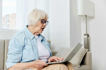 an elderly woman, passionate about work, is sitting in a bright cozy room in a stylish shirt, working on a laptop from home, typing text