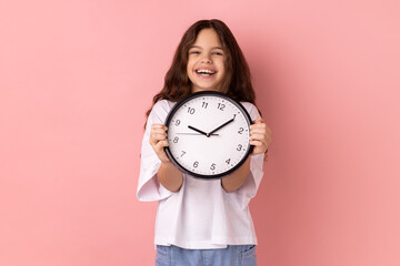 Portrait of smiling delighted little girl wearing white T-shirt holding wall clock, being happy,...