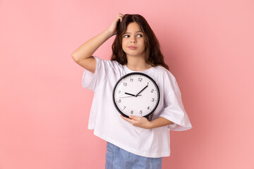 Portrait of confused puzzled little girl wearing white T-shirt holding wall clock and scratching...