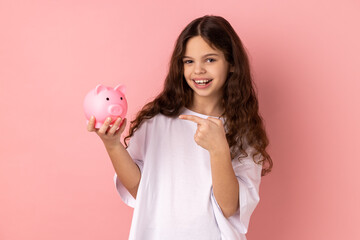 Portrait of smiling little girl wearing white T-shirt pointing at pink piggy bank, looking at...